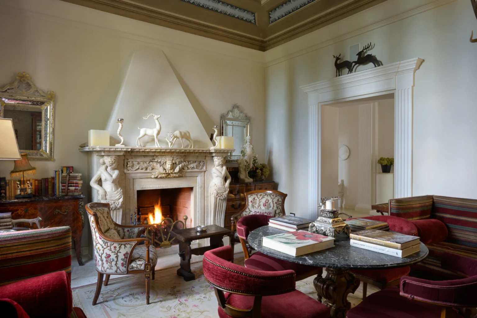 Elegant living room featuring a marble fireplace adorned with decorative sculptures, plush seating, and a round marble table surrounded by rich red chairs. Natural light enhances the sophisticated ambiance of the space, complemented by ornate details and a cozy fire. Ideal for showcasing luxury interior design and home decor inspiration. | Only on SopranoVillas