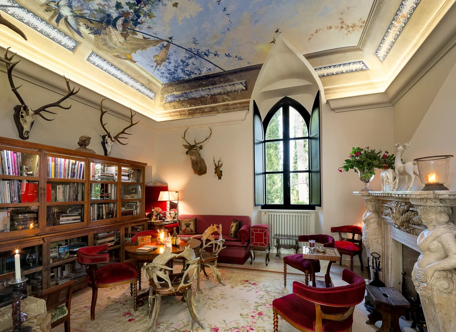 Elegant interior of a cozy living room featuring a beautifully painted ceiling, vintage furniture, and decorative antler mounts. The space includes a wooden bookshelf filled with books, a plush red sofa, and a marble fireplace adorned with floral arrangements, creating a warm and inviting atmosphere. | Only on SopranoVillas