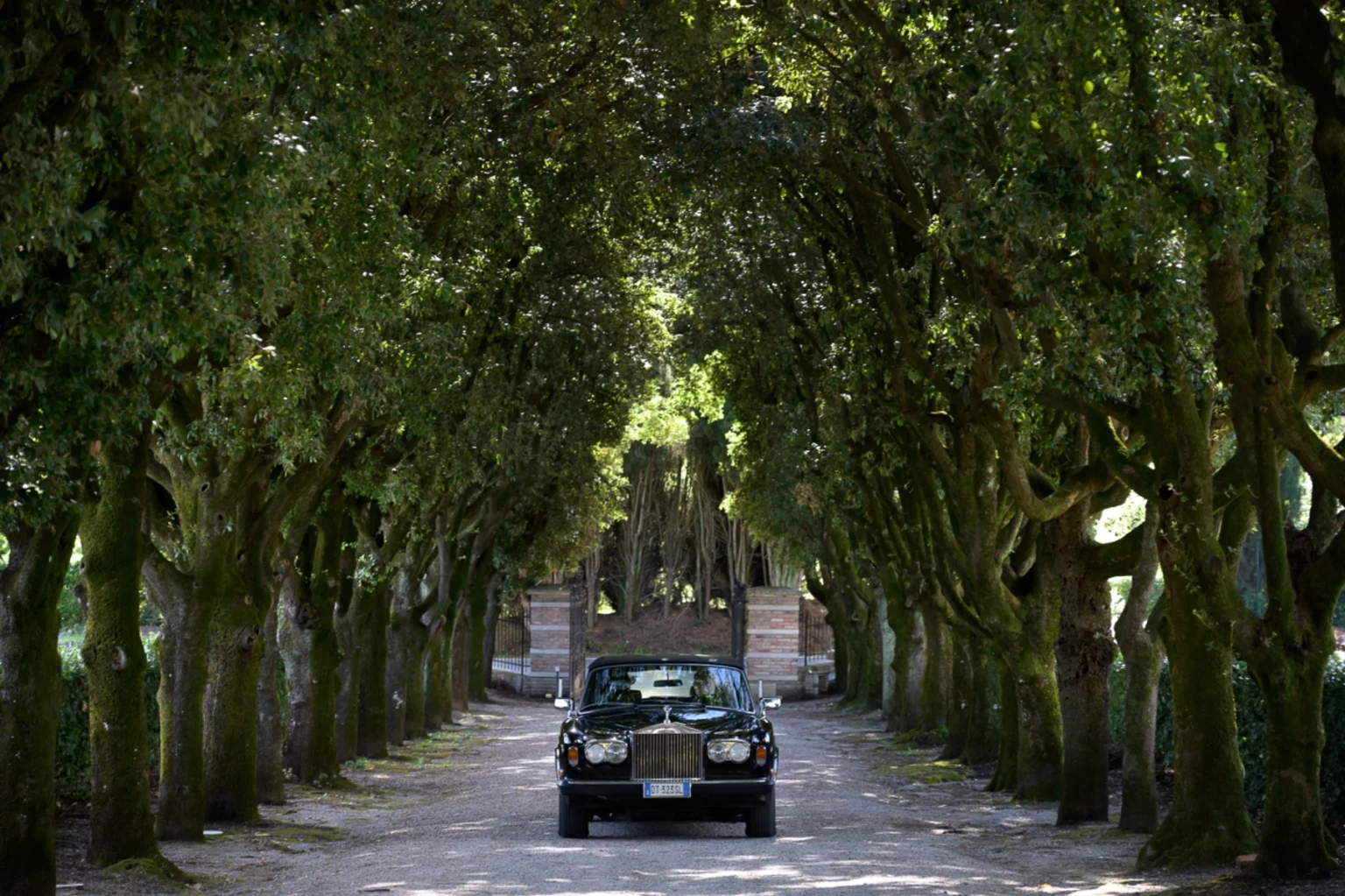 Black Rolls-Royce parked on a gravel driveway lined with lush, green trees, leading to a gated entrance. The sunlight filters through the foliage, creating a serene and elegant atmosphere. | Only on SopranoVillas