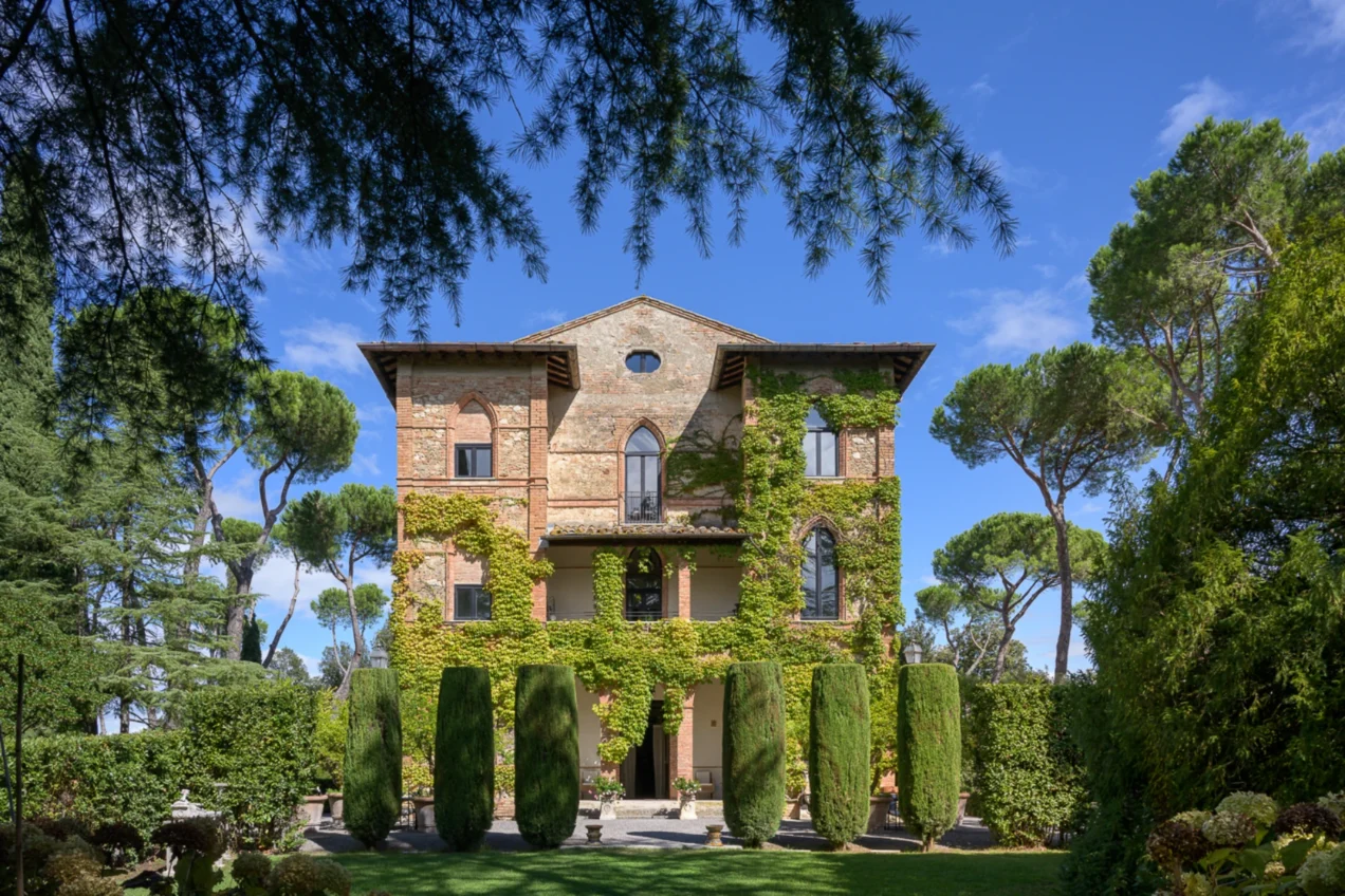 Historic villa covered in greenery, featuring stone architecture and tall cypress trees in a lush garden setting under a clear blue sky. | Only on SopranoVillas