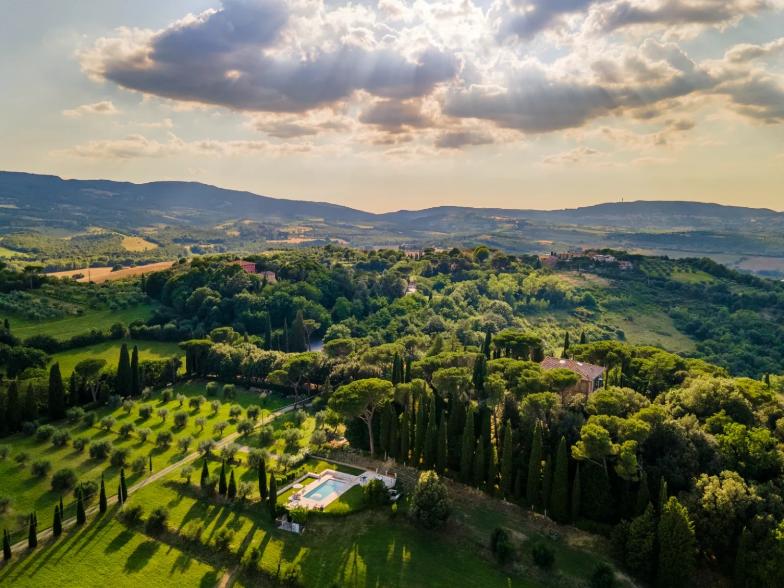 Aerial view of a serene Tuscan landscape featuring lush green hills, rows of cypress trees, and a private swimming pool surrounded by olive groves, under partly cloudy skies. | Only on SopranoVillas
