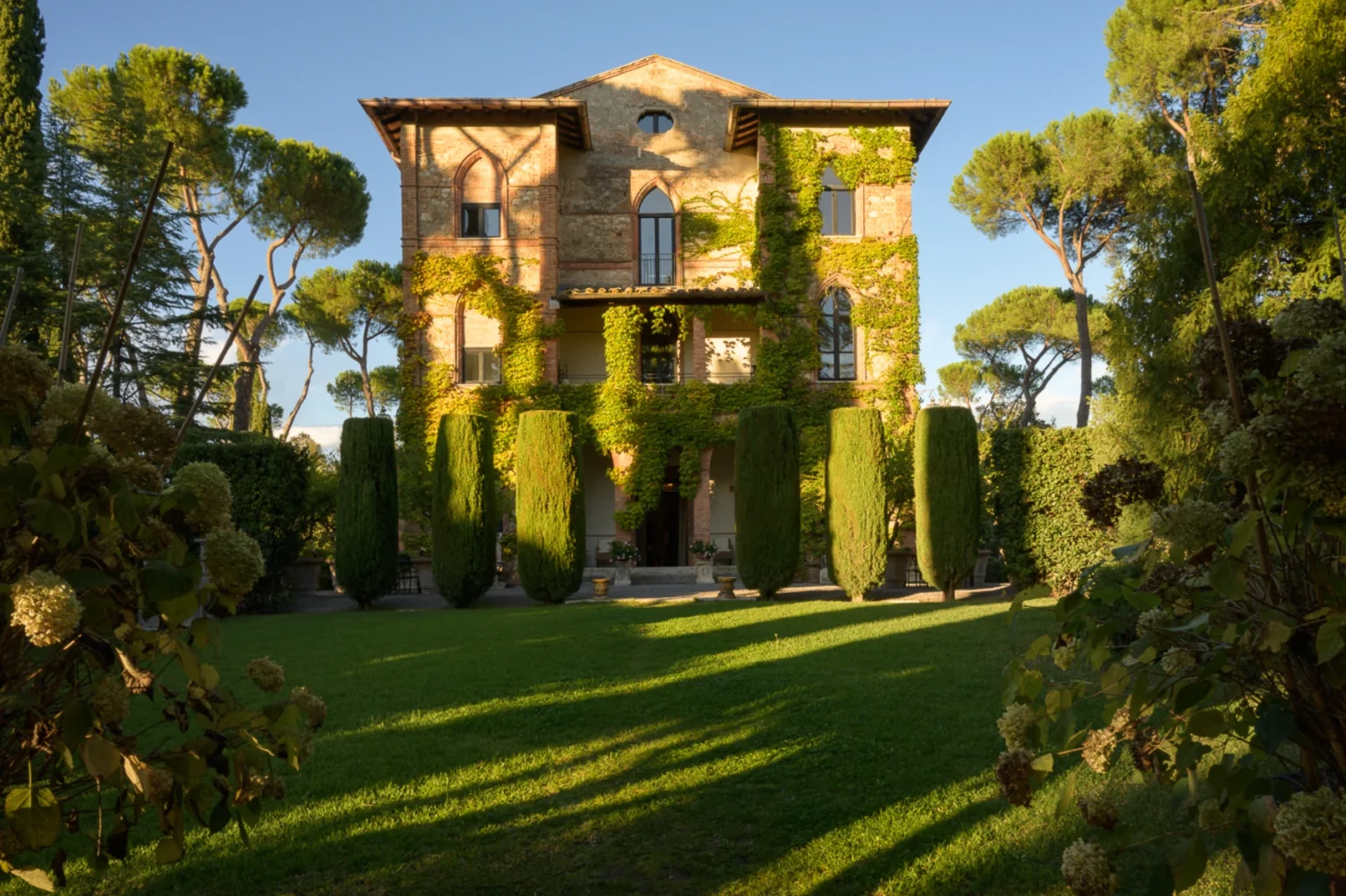 Elegant historic villa covered in greenery, surrounded by manicured hedges and tall cypress trees, set against a clear blue sky. The lush garden and architectural details highlight the beauty of this serene outdoor space. | Only on SopranoVillas