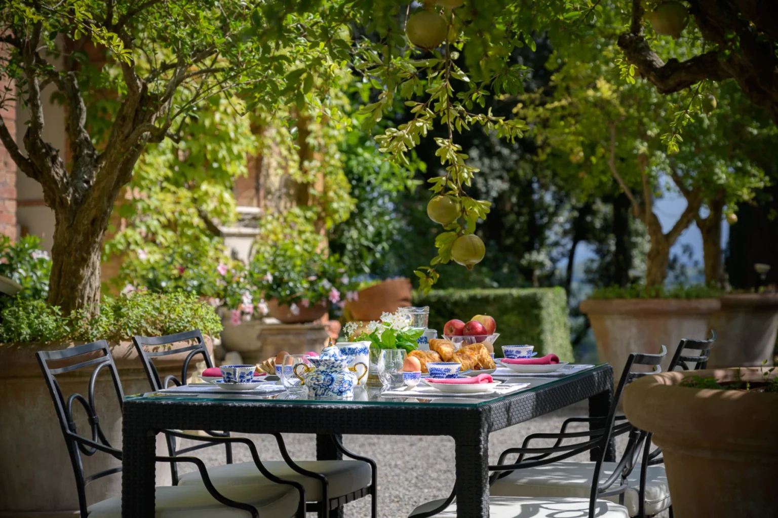 Outdoor dining setup featuring a beautifully arranged table with blue and white china, fresh fruits, pastries, and flowers, surrounded by lush greenery and trees in a serene garden setting. | Only on SopranoVillas
