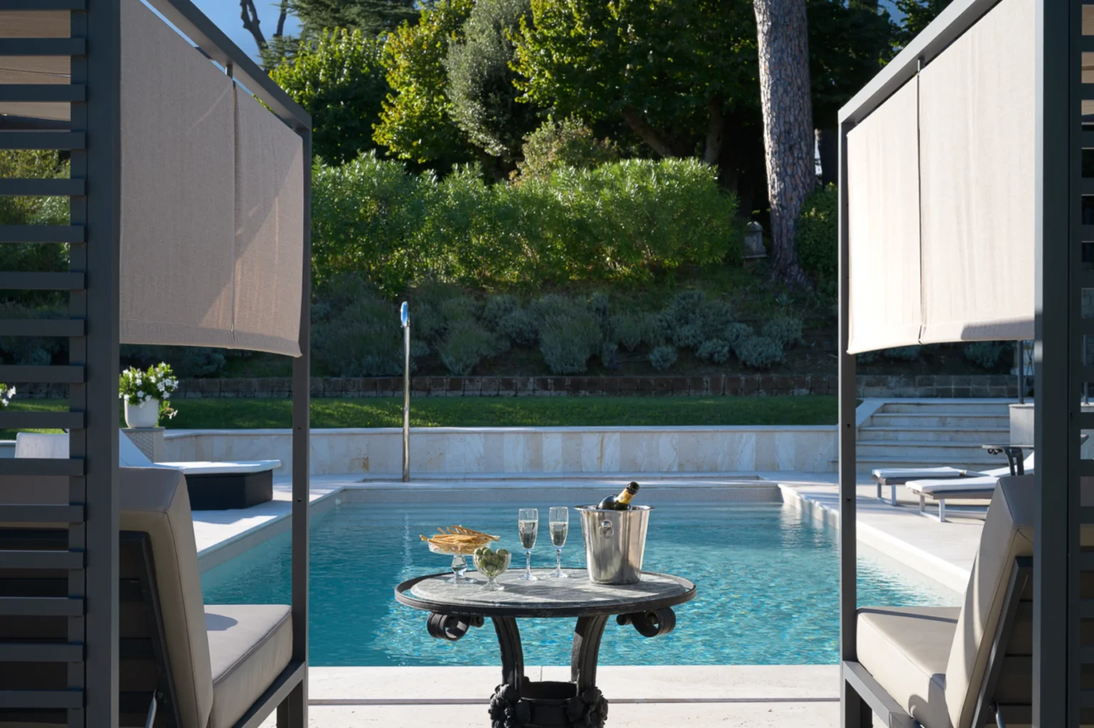 Luxurious outdoor poolside setting featuring a table with a champagne bucket, two flutes, and a plate of snacks, framed by elegant seating and greenery in the background. | Only on SopranoVillas