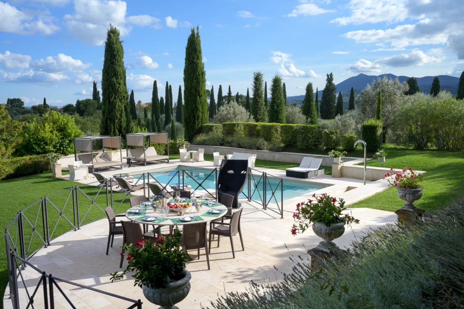 Luxurious outdoor patio with a dining table set for a meal, surrounded by lush greenery and cypress trees, featuring a swimming pool and comfortable lounge chairs under a clear blue sky. | Only on SopranoVillas