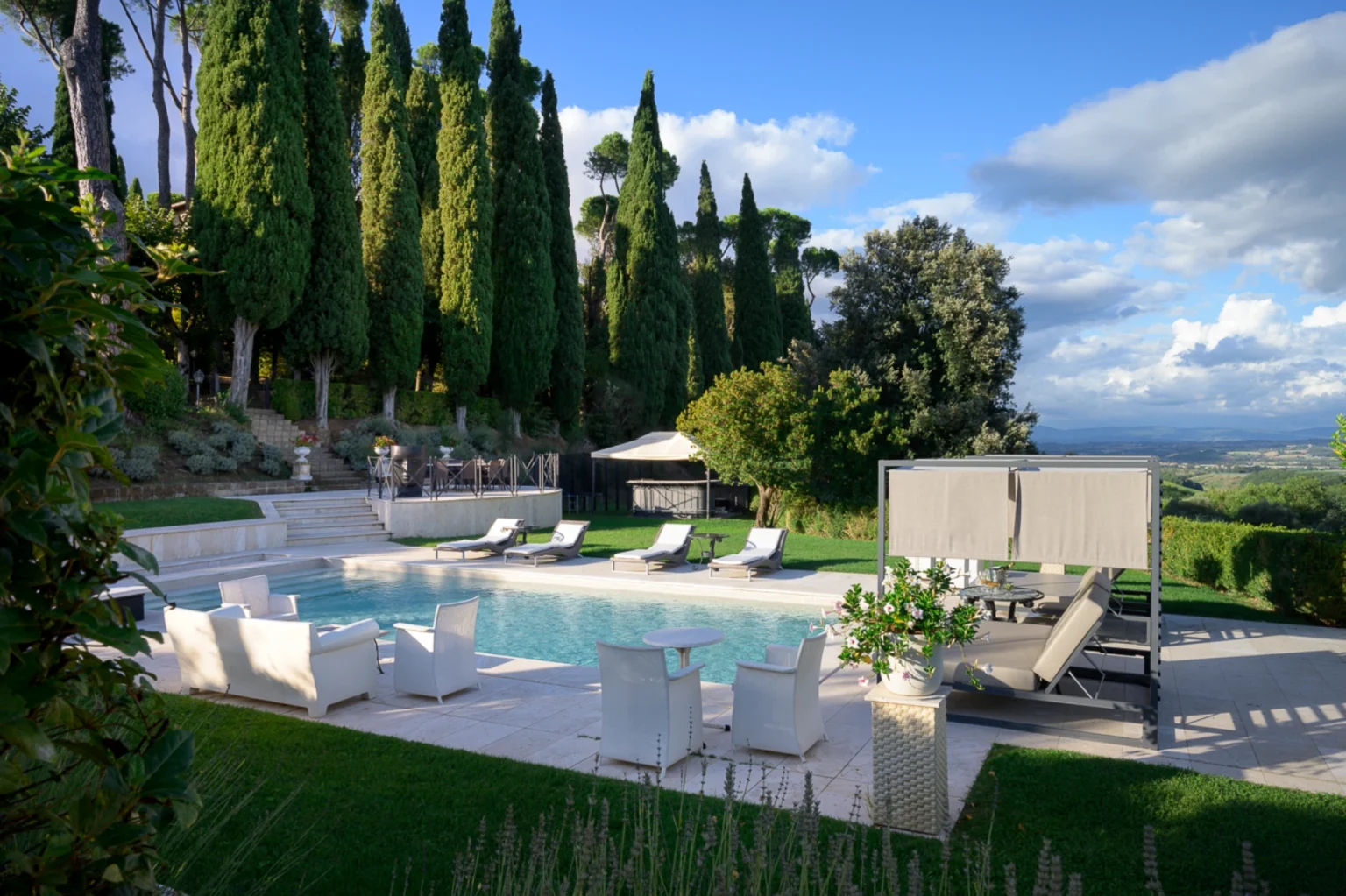 Luxurious outdoor pool area featuring modern lounge chairs, white seating, and lush greenery, surrounded by tall cypress trees and a scenic view. Ideal for relaxation and entertainment in a serene setting. | Only on SopranoVillas