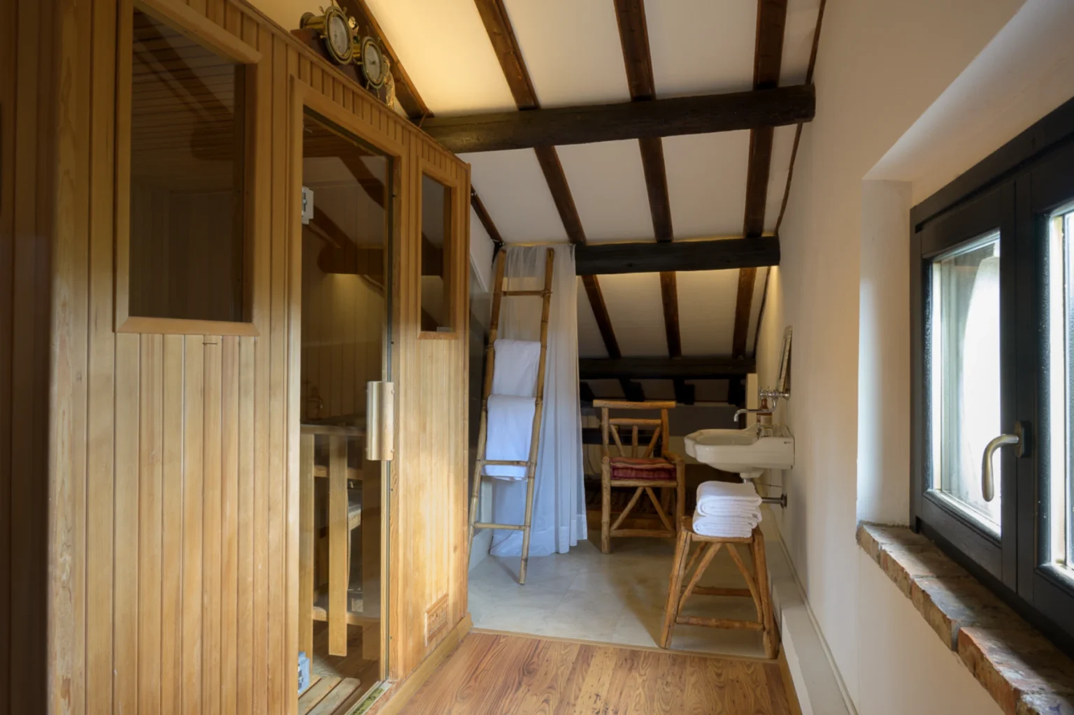 Cozy attic spa room featuring a wooden sauna, a ladder with towels, a rustic chair, and a modern sink, accented by exposed wooden beams and natural light. Perfect for relaxation and wellness. | Only on SopranoVillas
