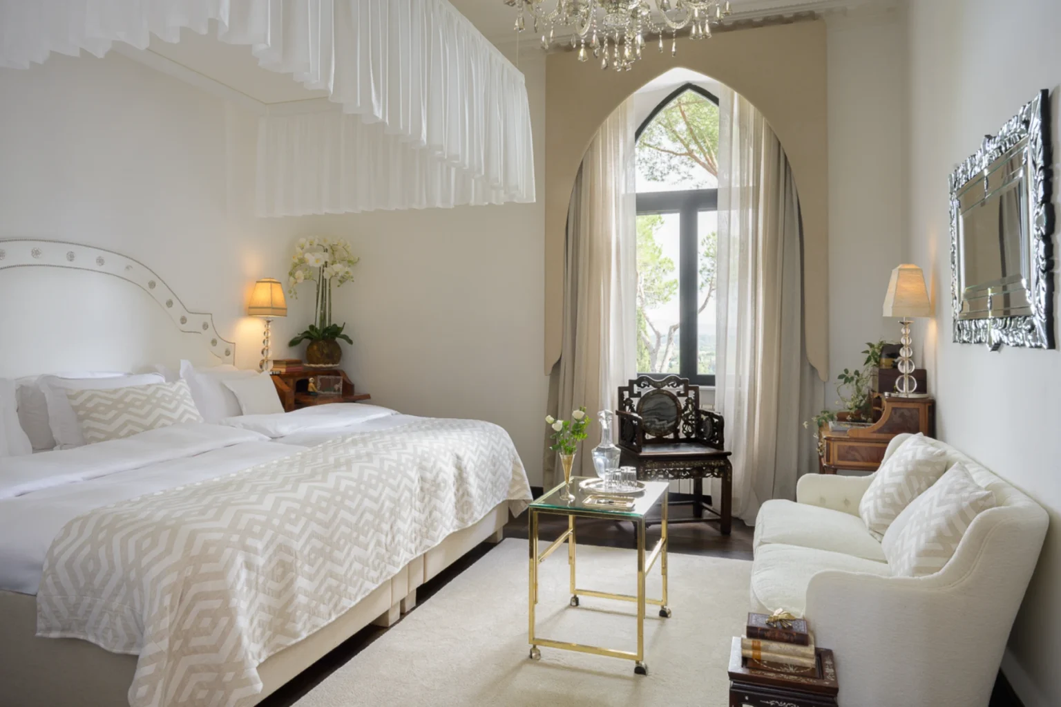 Elegant hotel room with a luxurious king-size bed, featuring a patterned throw blanket and decorative pillows. The room includes a stylish seating area with a white sofa, a glass coffee table, and an ornate chair. Natural light streams through large windows adorned with sheer curtains, complementing the chic decor highlighted by a chandelier and potted orchids. Perfect for a relaxing getaway or romantic retreat. | Only on SopranoVillas