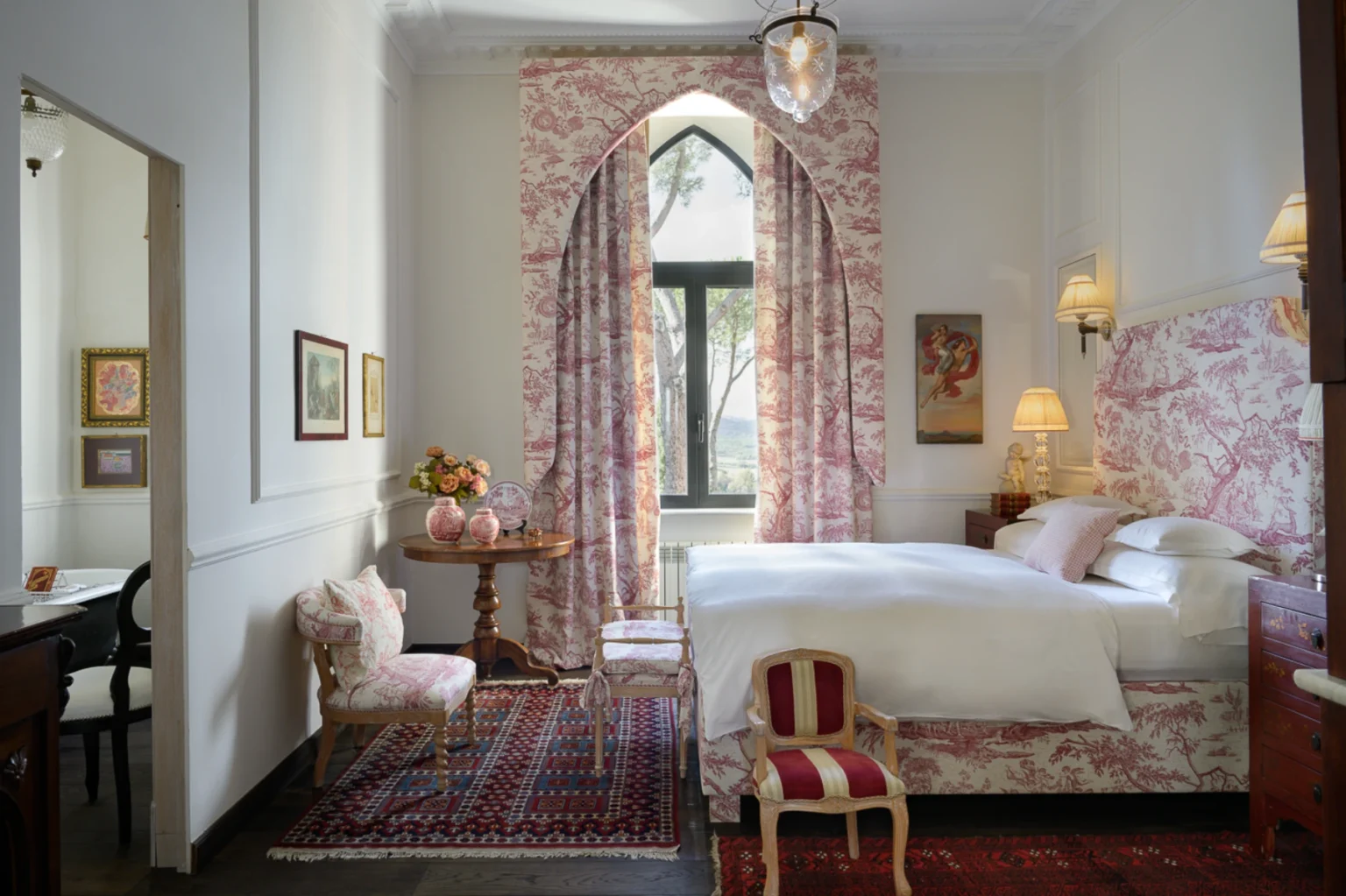 Elegant bedroom featuring floral-patterned curtains and bed linens, a wooden table with floral arrangements, and vintage decor. The room is illuminated by stylish lamps and showcases a cozy seating area with a plush chair, all set against a backdrop of large windows offering natural light. | Only on SopranoVillas