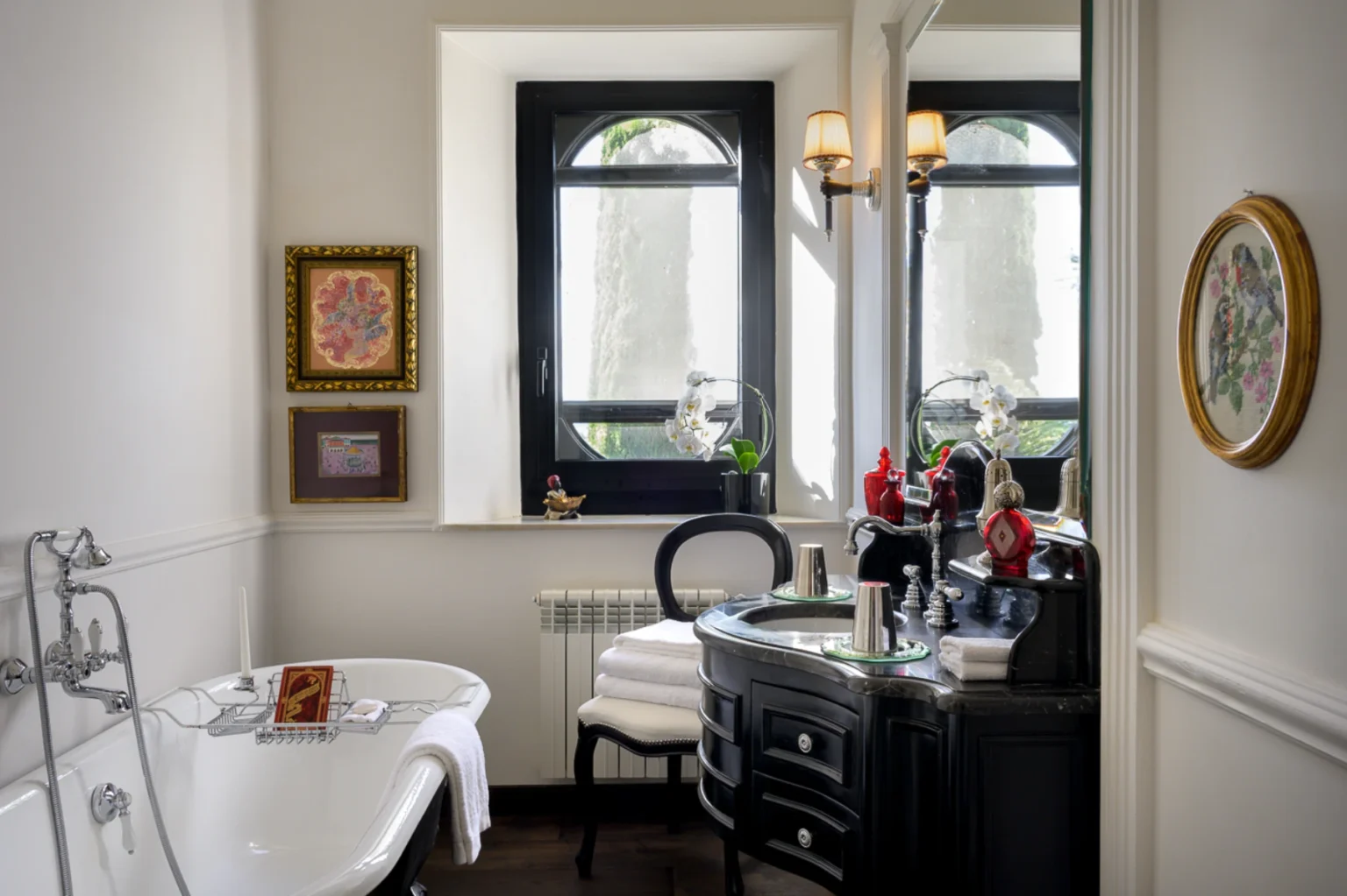 Elegant bathroom design featuring a freestanding bathtub, stylish black vanity with decorative accessories, framed artwork on the walls, and natural light streaming through large windows. | Only on SopranoVillas