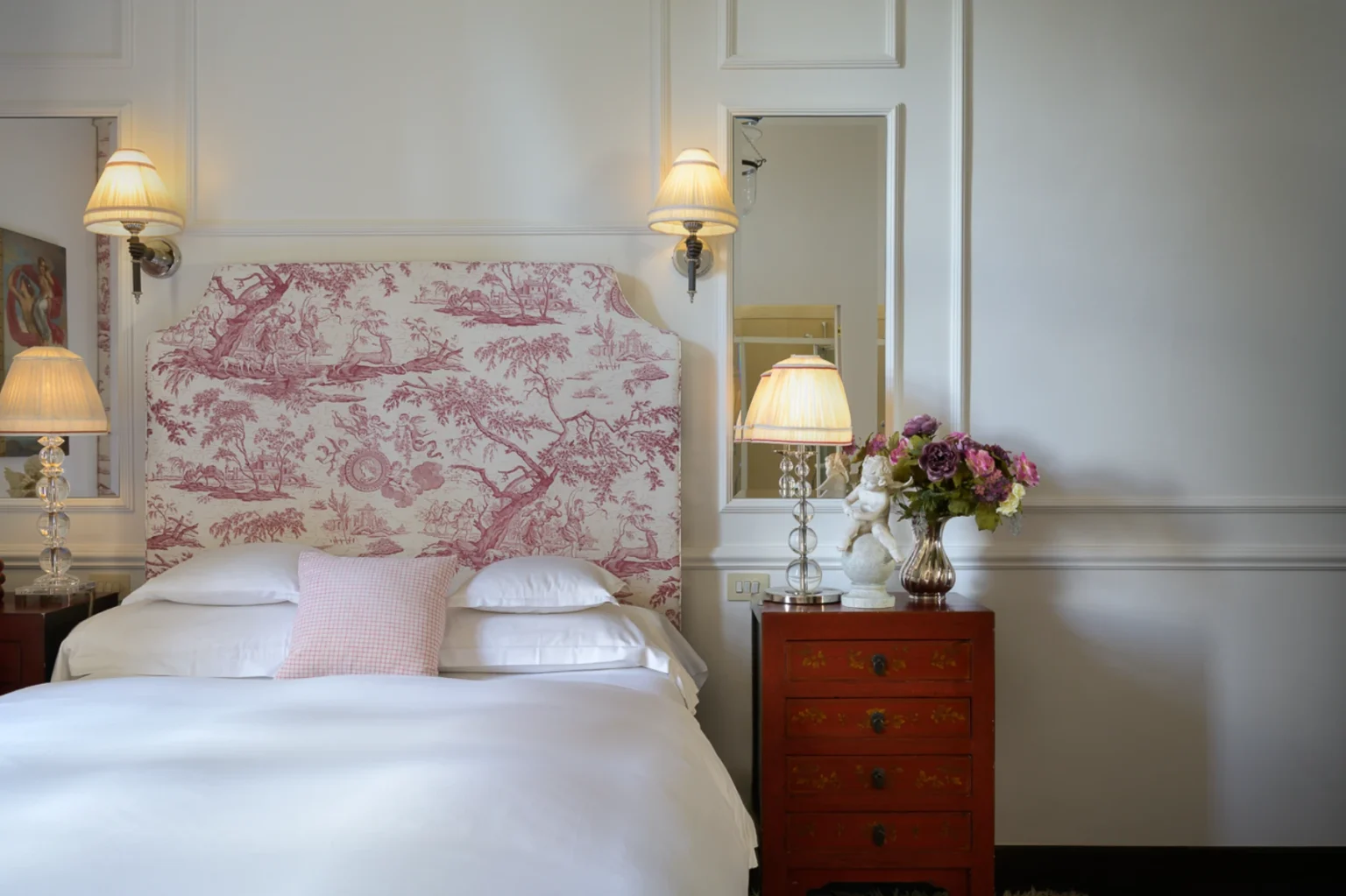 Cozy bedroom featuring a vintage pink and white toile headboard, elegant bedside lamps, and a decorative red nightstand adorned with flowers and a statue. The soft white bedding and neutral wall colors create a serene atmosphere. | Only on SopranoVillas