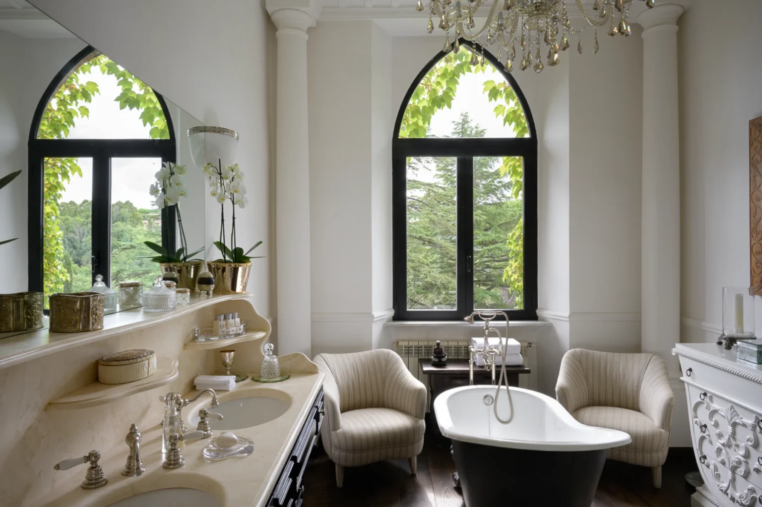 Luxurious bathroom featuring a freestanding black and white bathtub, elegant chandelier, and arched windows with greenery outside. The vanity area includes stylish decor, polished fixtures, and potted orchids, creating a serene and sophisticated atmosphere. | Only on SopranoVillas