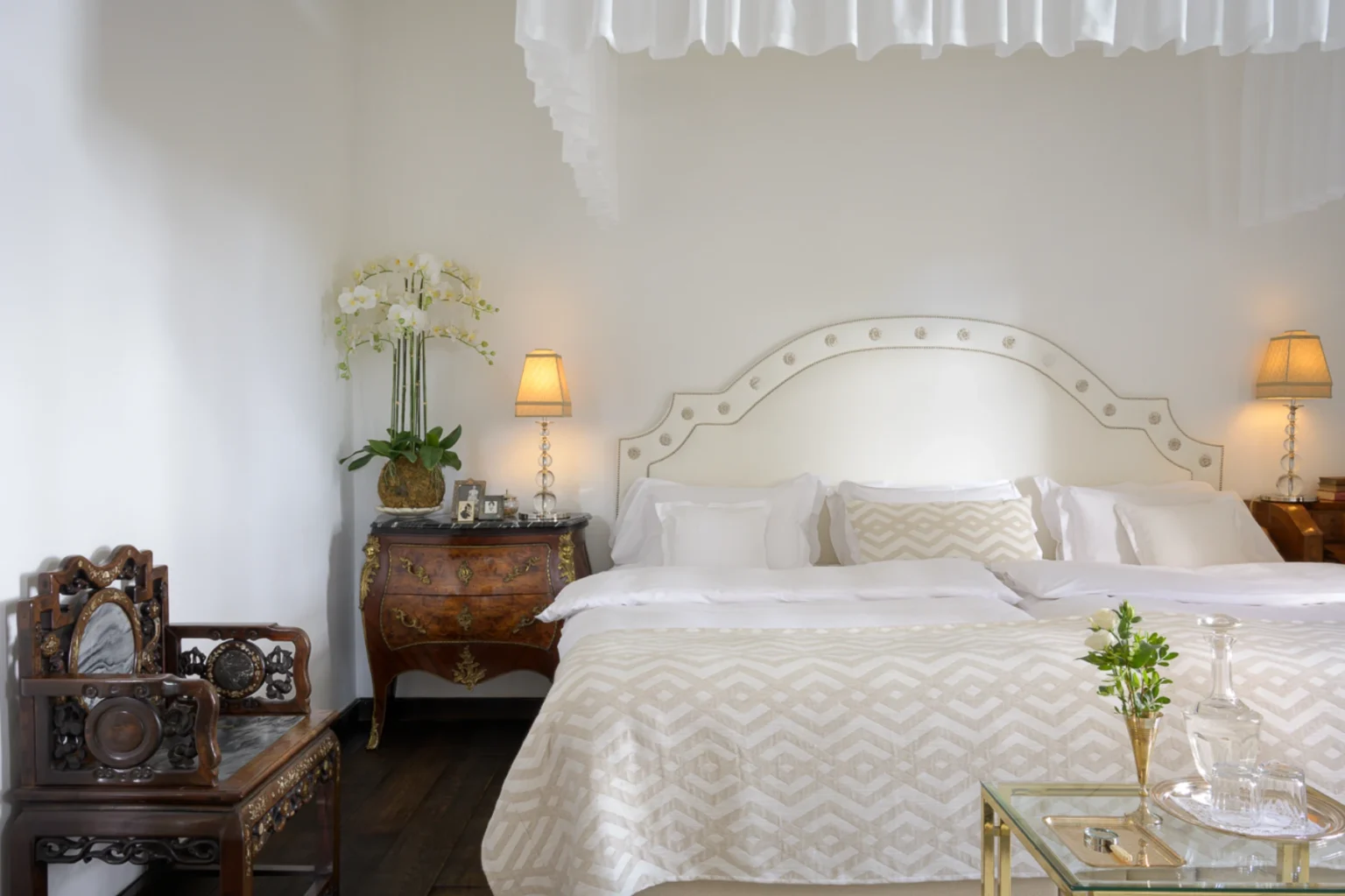 Elegant bedroom featuring a plush king-size bed with decorative pillows, a vintage wooden chair, and stylish bedside lamps. The room is adorned with a vase of orchids and a glass side table, creating a serene and luxurious atmosphere. Ideal for showcasing modern interior design and comfort. | Only on SopranoVillas