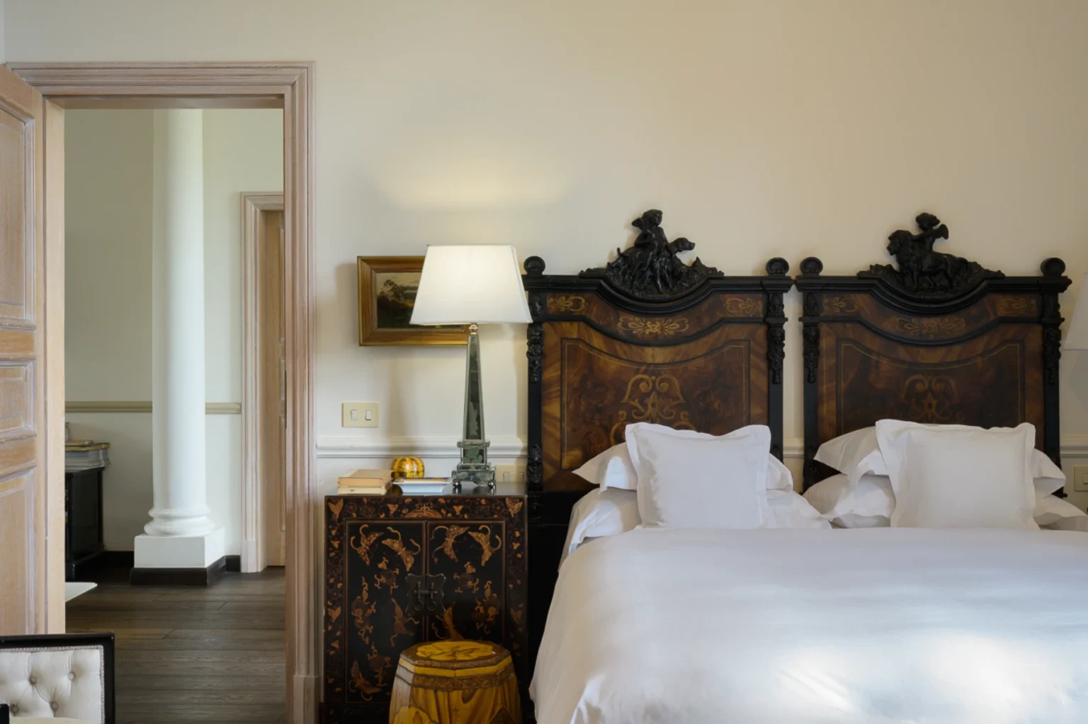 Elegant bedroom featuring intricately designed wooden headboards, a stylish bedside table with a lamp, and plush white bedding. The room is well-lit, showcasing a warm color palette and classic decor elements. | Only on SopranoVillas