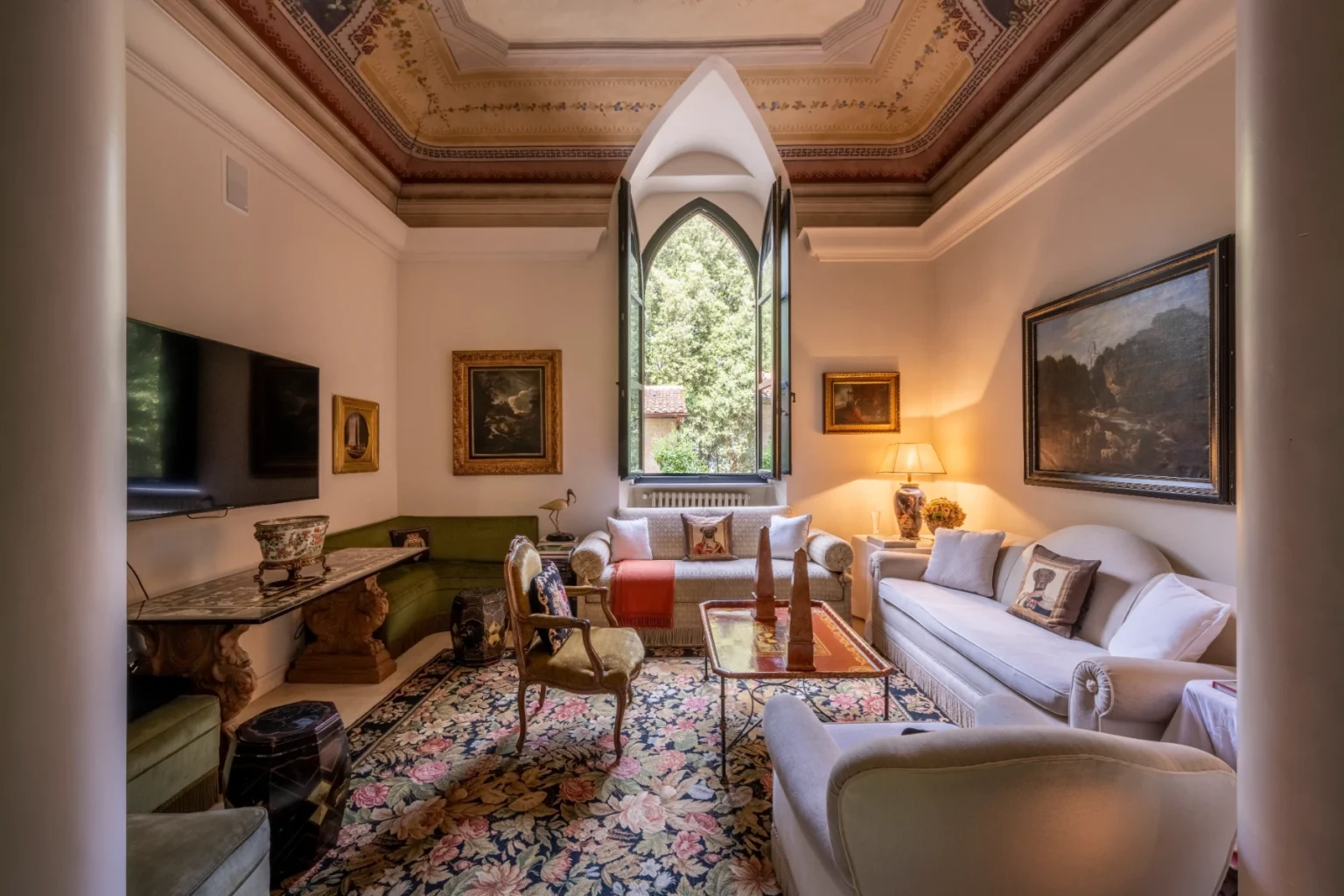 Elegant living room with vintage decor, featuring a large window, plush seating, and ornate ceiling details. The space includes a green sofa, a stylish coffee table, and various artworks on the walls, creating a cozy and inviting atmosphere. | Only on SopranoVillas