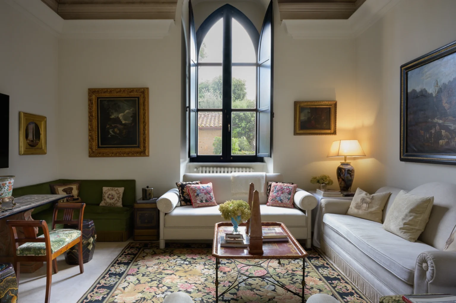 Elegant living room featuring a combination of classic and contemporary decor, including a white sofa with decorative pillows, a green velvet bench, and a wooden table. The room is illuminated by natural light from a large arched window, showcasing lush outdoor greenery. Artwork adorns the walls, enhancing the sophisticated atmosphere. A patterned area rug adds warmth to the space. | Only on SopranoVillas