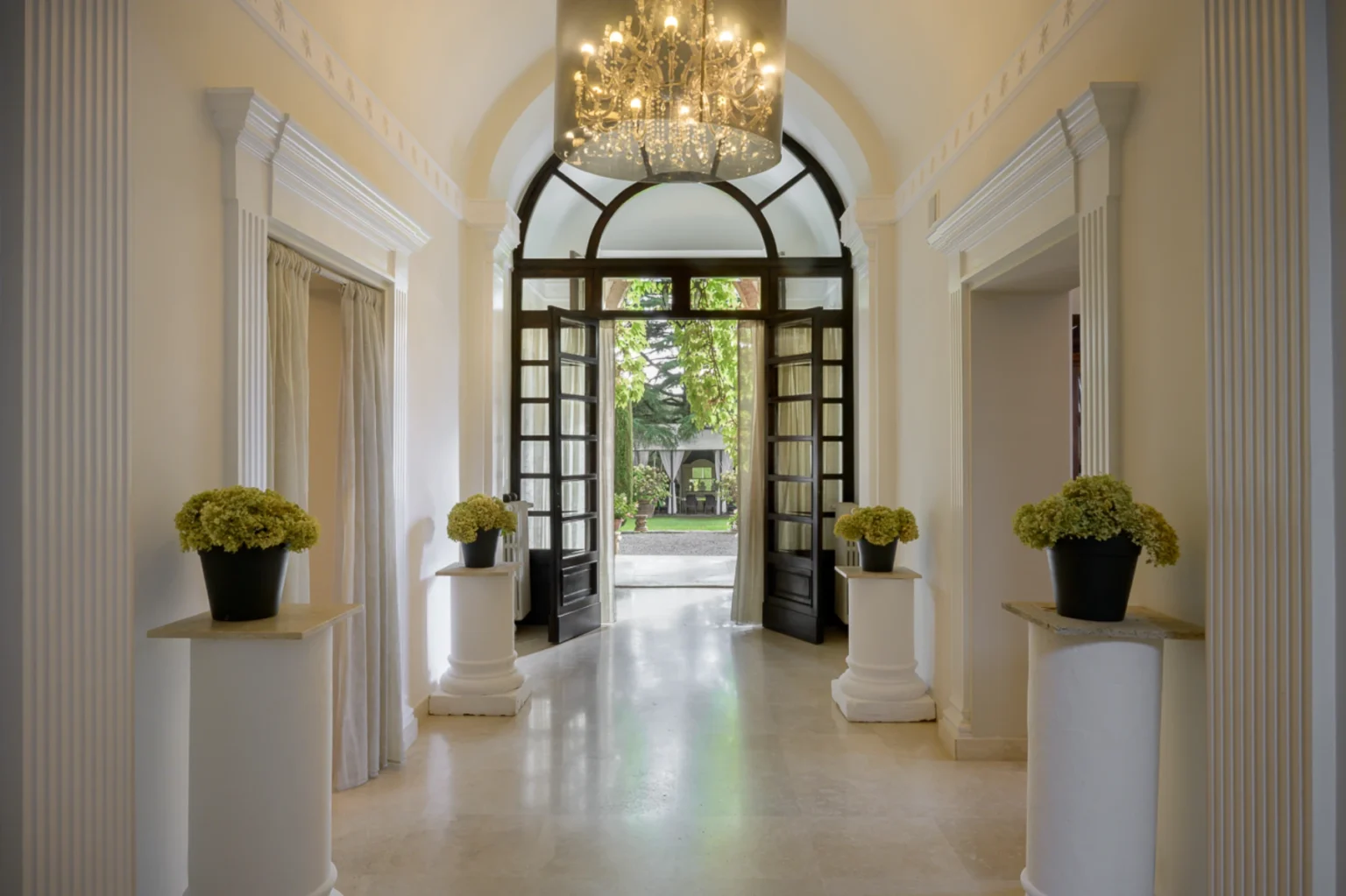 Elegant entrance hallway featuring open double doors, a chandelier, and potted green flowers on white pedestals, leading to a garden view. | Only on SopranoVillas