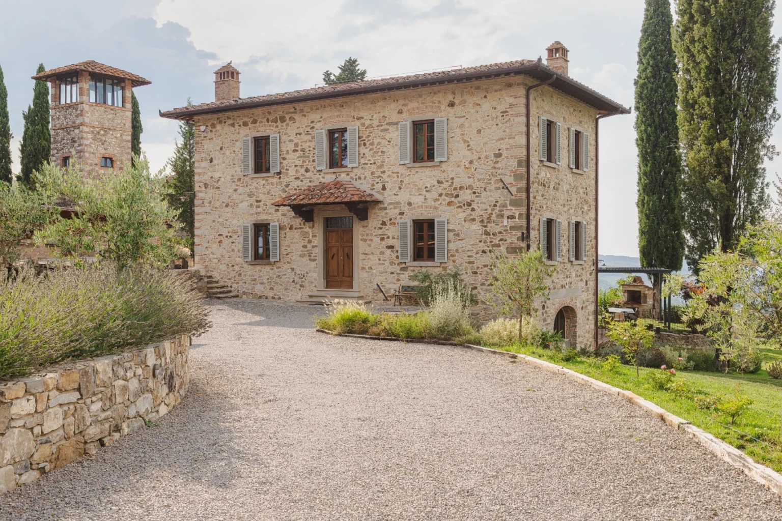 Charming rustic stone villa with a gravel driveway, surrounded by lush greenery and cypress trees, featuring a traditional tile roof and wooden shutters, set against a picturesque landscape. | Only on SopranoVillas