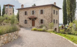 Charming rustic stone villa with a gravel driveway, surrounded by lush greenery and cypress trees, featuring a traditional tile roof and wooden shutters, set against a picturesque landscape.