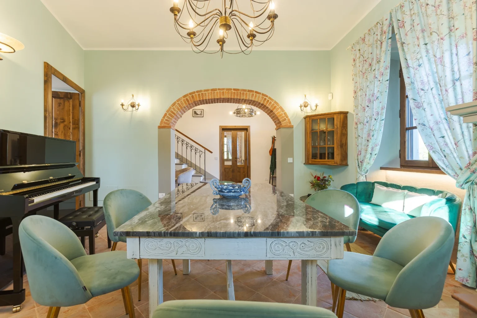 Modern dining area featuring a marble table, plush green chairs, and a grand piano, with decorative light fixtures and floral curtains, creating an inviting atmosphere in a stylish home. | Only on SopranoVillas