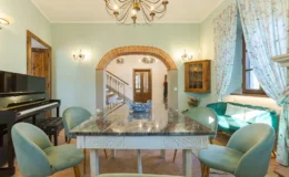 Modern dining area featuring a marble table, plush green chairs, and a grand piano, with decorative light fixtures and floral curtains, creating an inviting atmosphere in a stylish home.