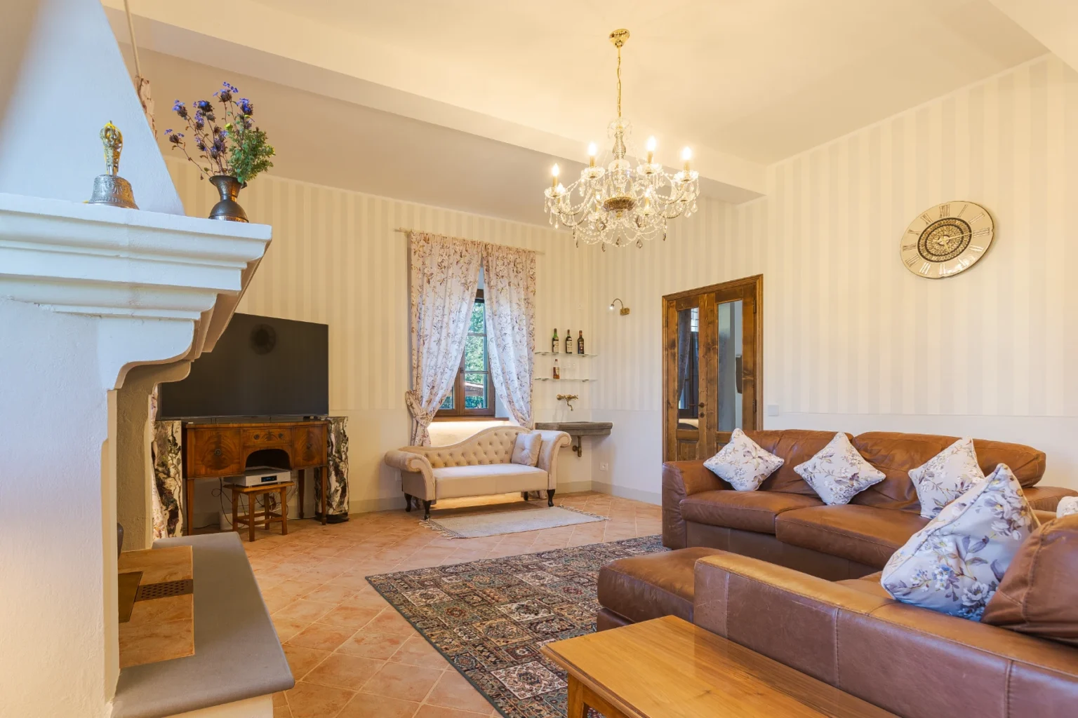 Cozy living room featuring a chandelier, leather sofas with decorative pillows, a vintage wooden console, and a flat-screen TV, all set against striped wallpaper and floral curtains, creating an inviting atmosphere. | Only on SopranoVillas