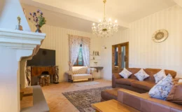 Cozy living room featuring a chandelier, leather sofas with decorative pillows, a vintage wooden console, and a flat-screen TV, all set against striped wallpaper and floral curtains, creating an inviting atmosphere.