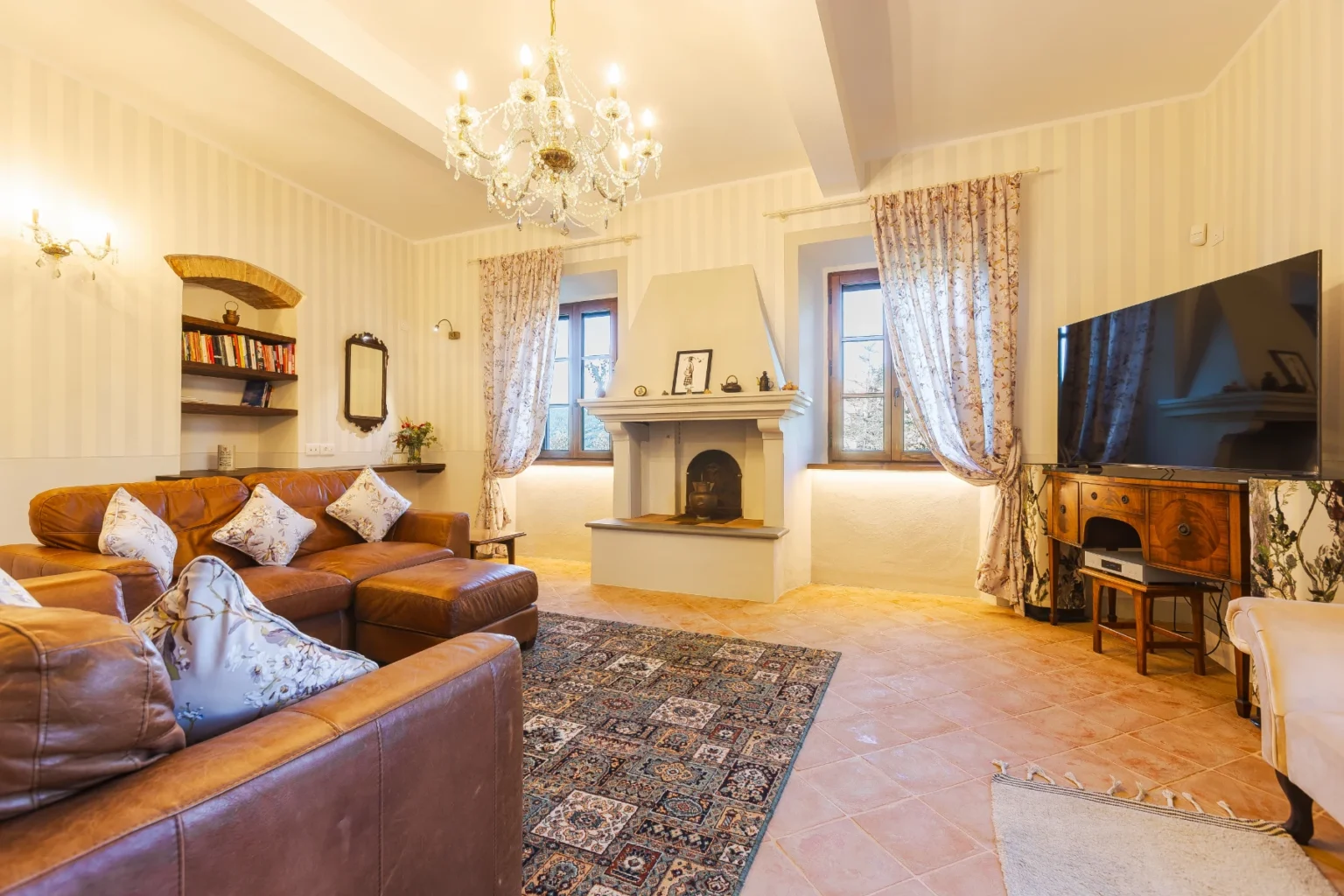 Cozy living room featuring leather sofas, a decorative chandelier, and a fireplace. The space includes large windows with floral curtains, a TV on a vintage-style cabinet, and a bookshelf filled with books, creating a warm and inviting atmosphere. | Only on SopranoVillas