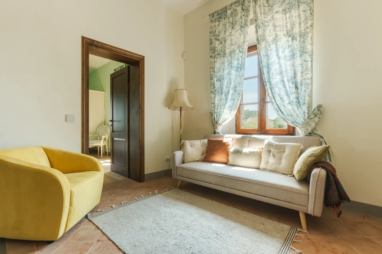 Cozy living room featuring a light gray sofa with decorative pillows, a vibrant yellow armchair, and a soft area rug. The room is illuminated by natural light from a window adorned with patterned curtains, and a stylish floor lamp adds to the decor. A doorway leads to another room with a green accent wall and a white chair. | Only on SopranoVillas