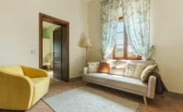 Cozy living room featuring a light gray sofa with decorative pillows, a vibrant yellow armchair, and a soft area rug. The room is illuminated by natural light from a window adorned with patterned curtains, and a stylish floor lamp adds to the decor. A doorway leads to another room with a green accent wall and a white chair.