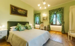 Cozy bedroom with green walls, a large bed adorned with decorative pillows, and elegant curtains. Features include a wooden floor, a stylish chandelier, and tasteful artwork on the walls, creating a serene and inviting atmosphere.