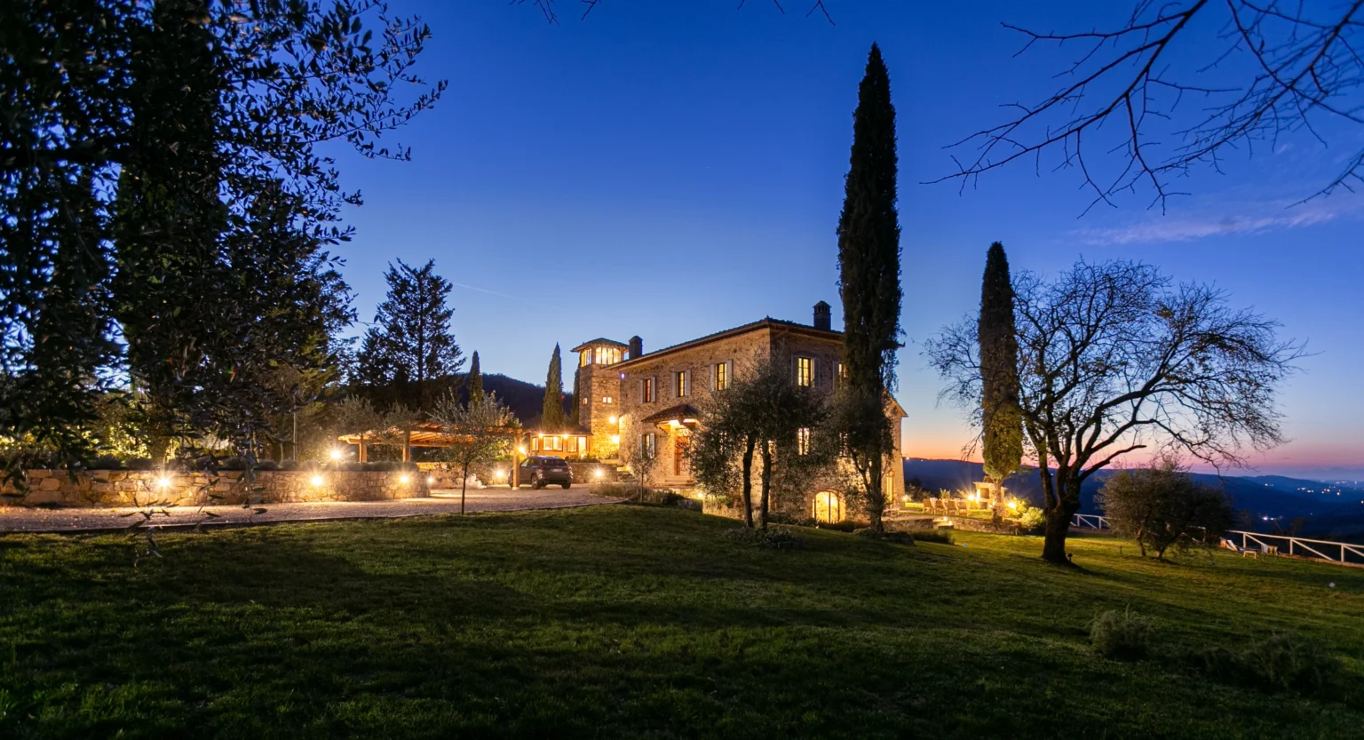 Beautiful stone villa illuminated at dusk, surrounded by lush greenery and tall cypress trees, set against a vibrant twilight sky. Perfect for luxury retreats or romantic getaways in a serene countryside setting. | Only on SopranoVillas