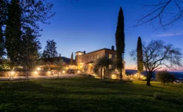 Beautiful stone villa illuminated at dusk, surrounded by lush greenery and tall cypress trees, set against a vibrant twilight sky. Perfect for luxury retreats or romantic getaways in a serene countryside setting.
