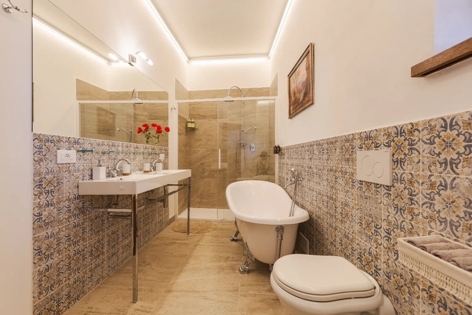 Modern bathroom interior featuring a freestanding bathtub, double sink vanity, and glass shower enclosure. Decorative patterned tiles add a stylish touch, complemented by natural light and fresh flowers. | Only on SopranoVillas