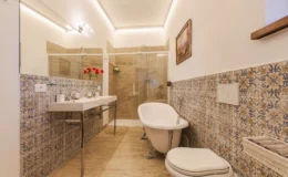 Modern bathroom interior featuring a freestanding bathtub, double sink vanity, and glass shower enclosure. Decorative patterned tiles add a stylish touch, complemented by natural light and fresh flowers.