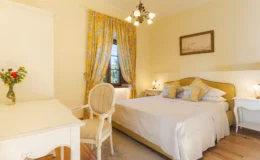 Cozy bedroom featuring a yellow and white color scheme, a queen-sized bed with decorative pillows, a vintage-style desk and chair, and elegant yellow curtains. The room is well-lit with natural light and includes a vase of fresh flowers, creating a warm and inviting atmosphere.