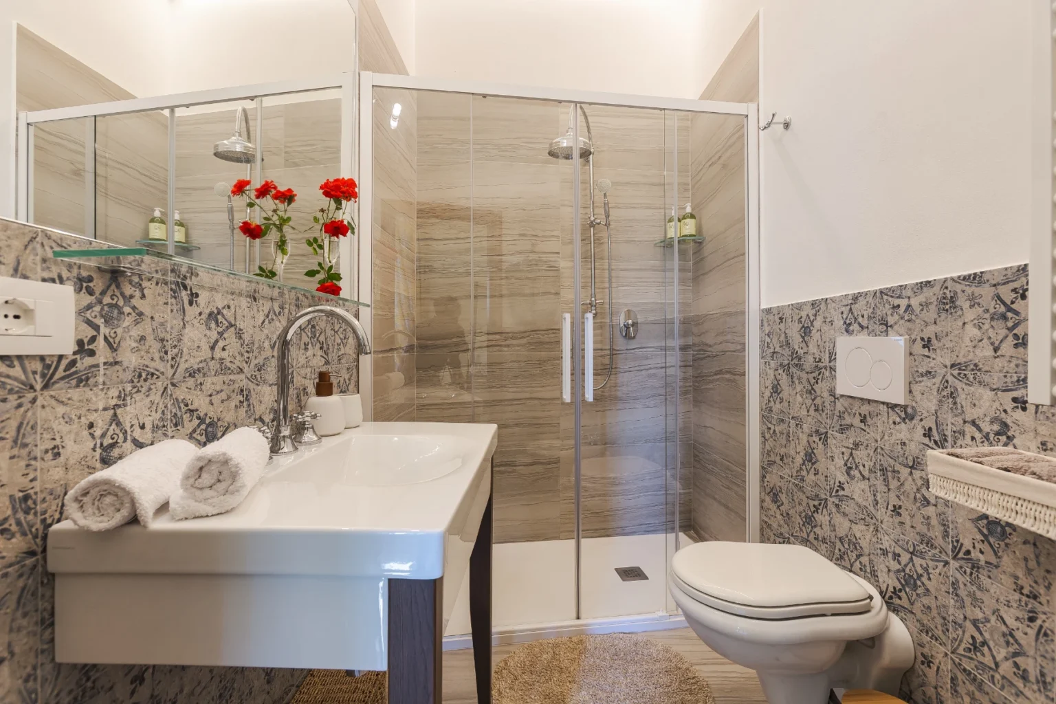 Modern bathroom featuring a glass shower enclosure, sleek sink with decorative towels, and stylish wall tiles. The space is enhanced by vibrant red flowers, creating a fresh and inviting atmosphere. | Only on SopranoVillas