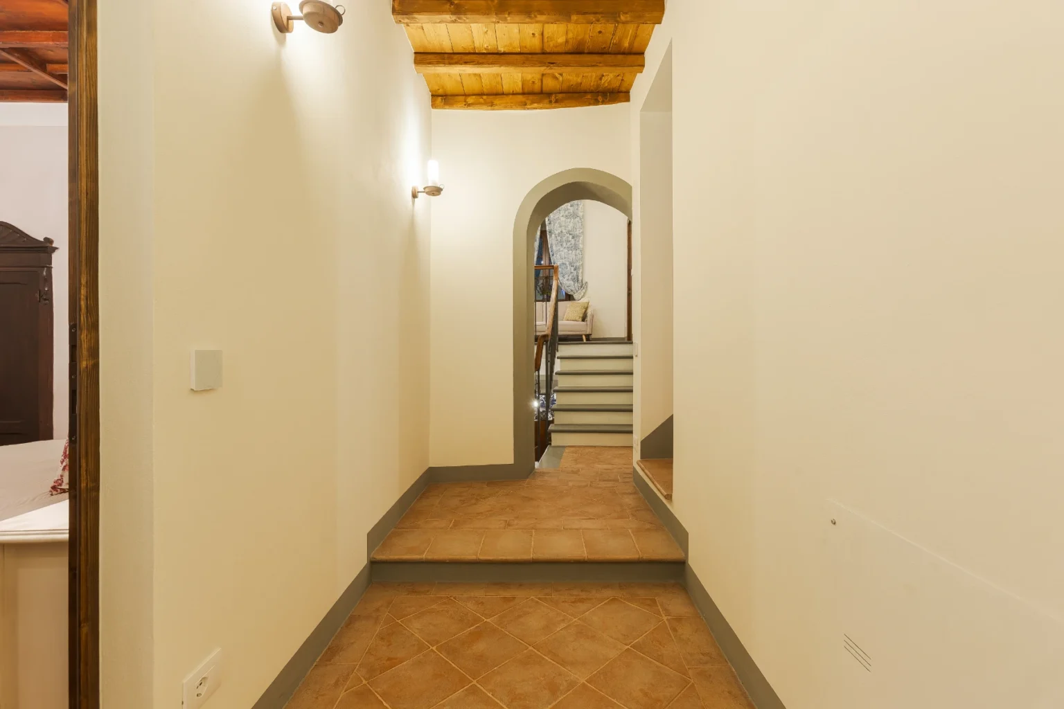 Interior hallway with a warm, inviting atmosphere, featuring terracotta tile flooring, white walls, and wooden ceiling beams. An arched doorway leads to a staircase, enhancing the home's spacious and elegant design. Soft lighting adds to the cozy ambiance. | Only on SopranoVillas
