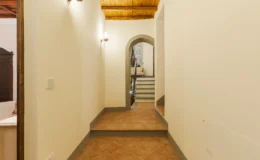 Interior hallway with a warm, inviting atmosphere, featuring terracotta tile flooring, white walls, and wooden ceiling beams. An arched doorway leads to a staircase, enhancing the home's spacious and elegant design. Soft lighting adds to the cozy ambiance.