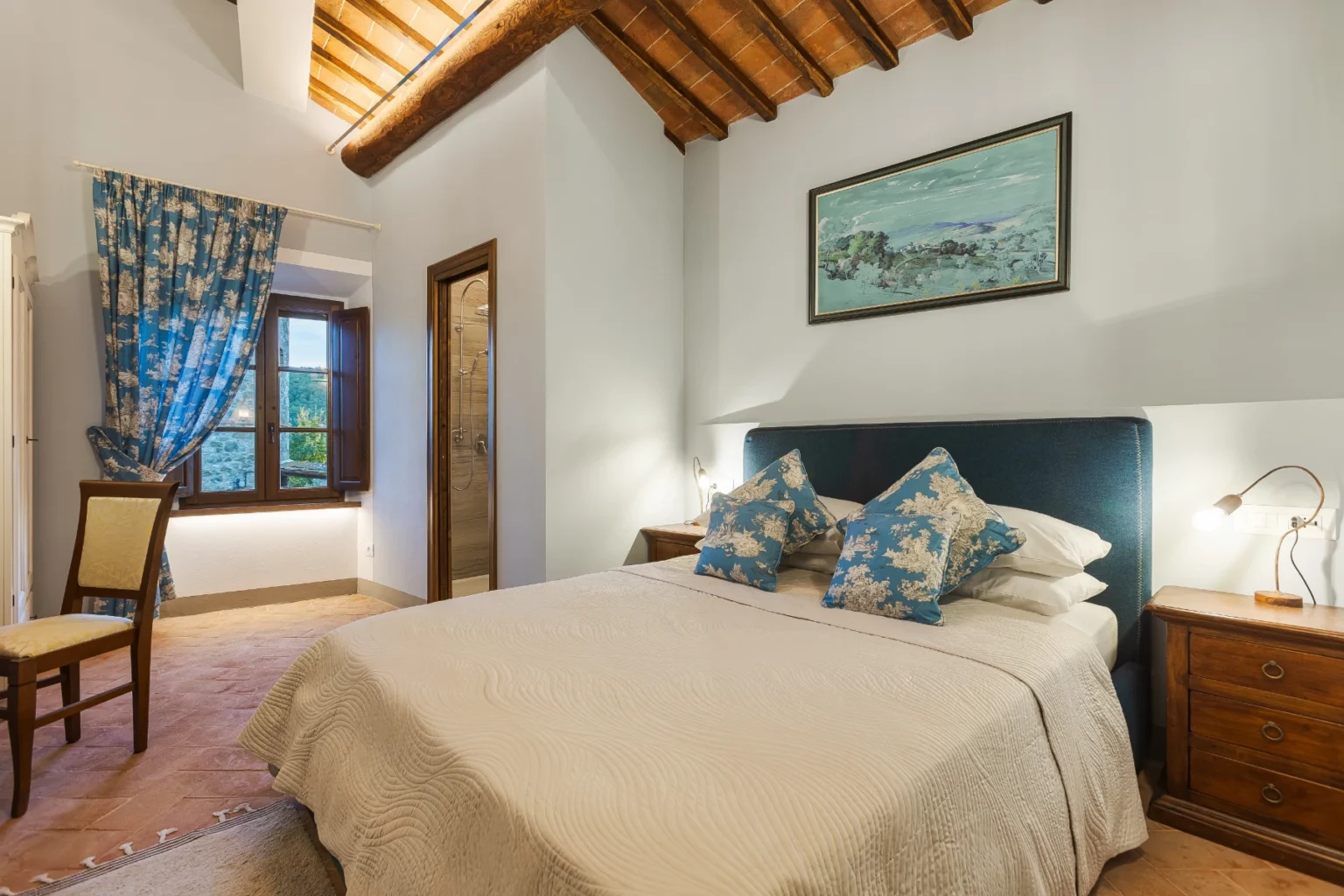 Cozy bedroom featuring a double bed with decorative blue pillows, wooden furniture, and a large window with floral curtains. The room includes a stylish en-suite bathroom and rustic ceiling beams, creating a warm and inviting atmosphere. Ideal for relaxation and comfort. | Only on SopranoVillas