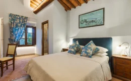Cozy bedroom featuring a double bed with decorative blue pillows, wooden furniture, and a large window with floral curtains. The room includes a stylish en-suite bathroom and rustic ceiling beams, creating a warm and inviting atmosphere. Ideal for relaxation and comfort.