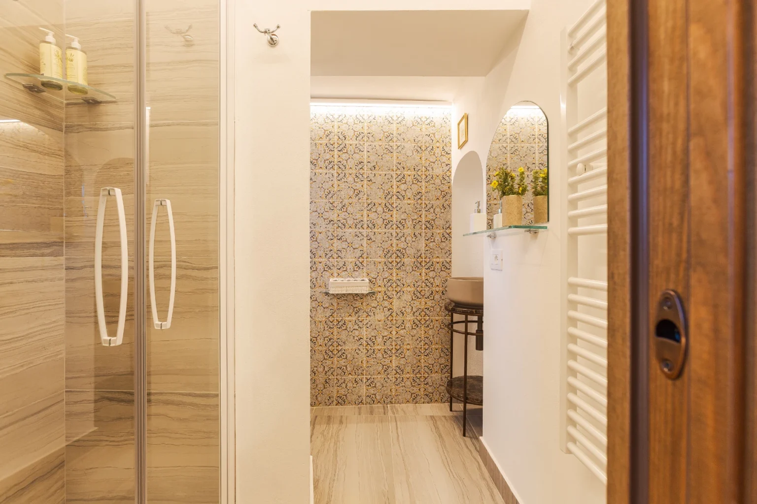 Modern bathroom interior featuring a glass shower enclosure, decorative tiled wall, and stylish fixtures. The space includes a small shelf with toiletries and a cozy seating area, complemented by natural light and contemporary design elements. | Only on SopranoVillas