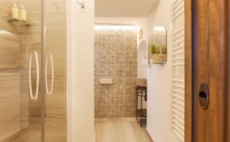 Modern bathroom interior featuring a glass shower enclosure, decorative tiled wall, and stylish fixtures. The space includes a small shelf with toiletries and a cozy seating area, complemented by natural light and contemporary design elements.