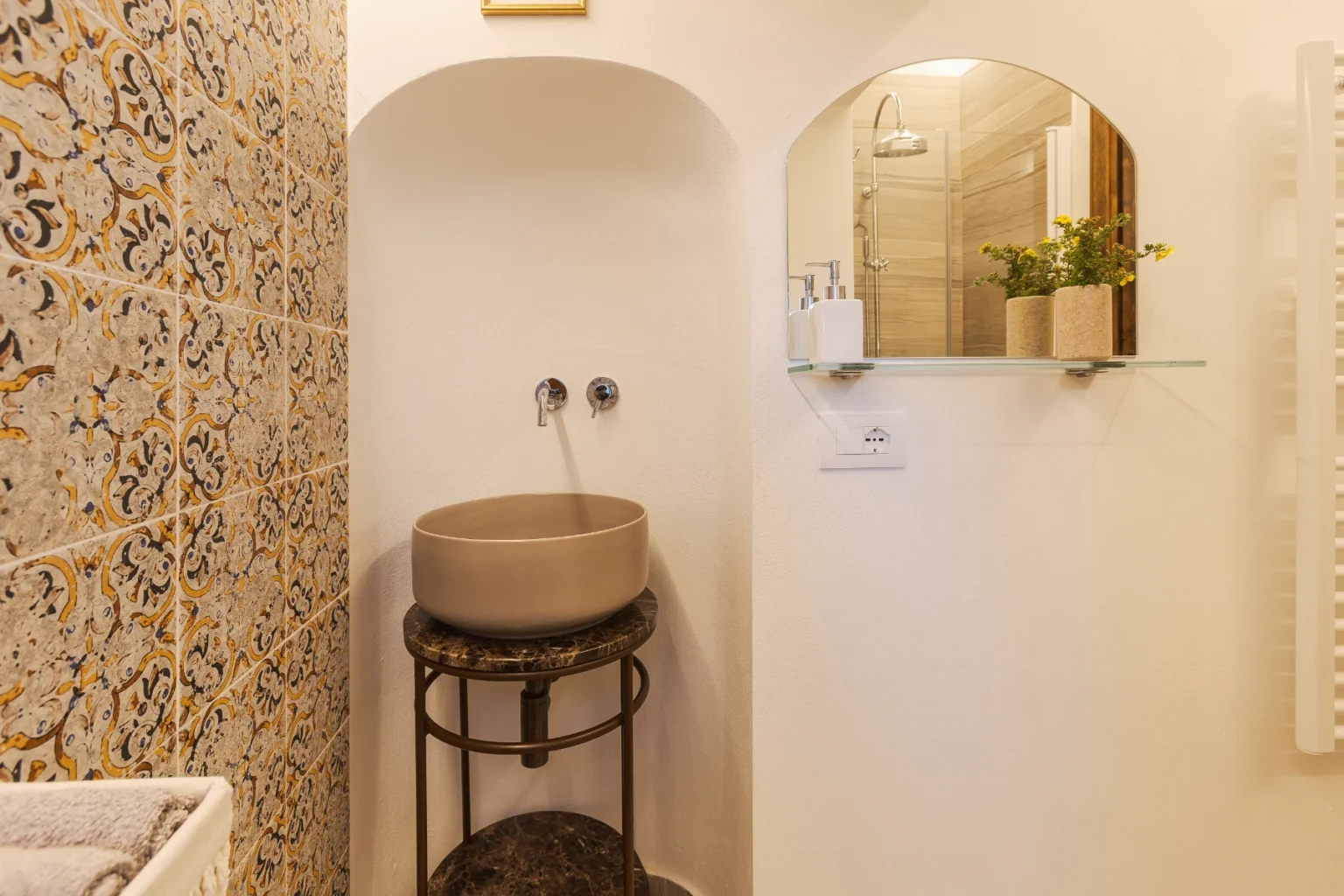 Modern bathroom design featuring a stylish round sink on a metal stand, decorative patterned tiles, a wall mirror, and minimalist shelving with potted plants. | Only on SopranoVillas