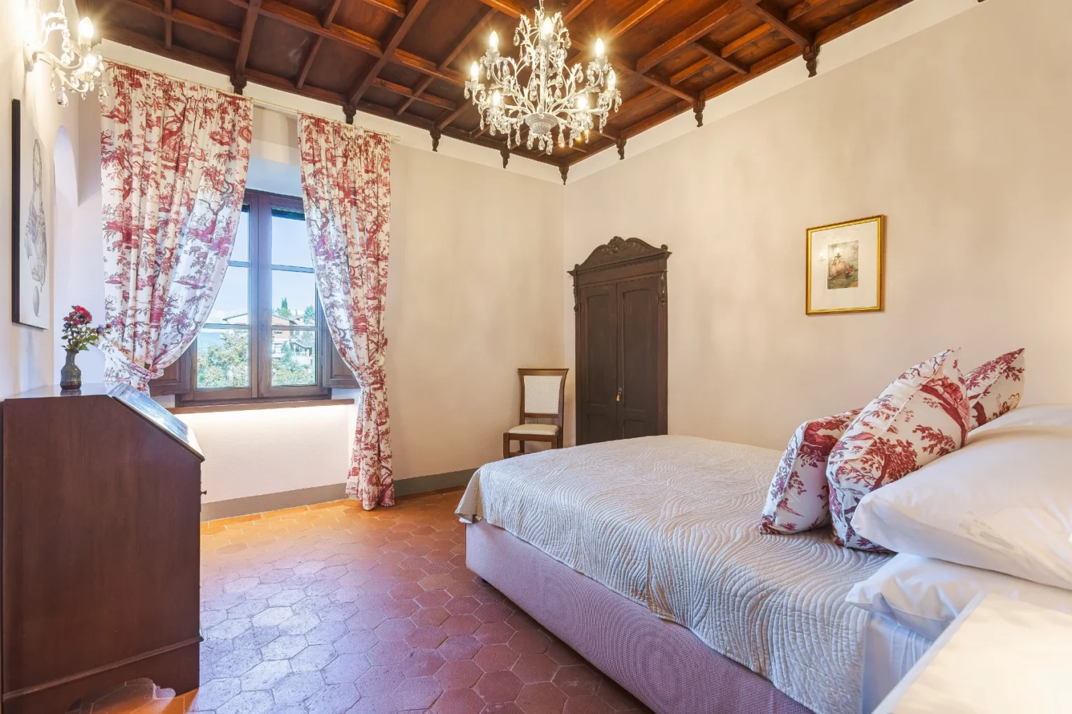 Cozy bedroom featuring a queen-sized bed with decorative pillows, elegant wooden furniture, and a vintage wardrobe. Natural light streams through large windows adorned with floral curtains, enhancing the inviting atmosphere. The room showcases a classic chandelier and terracotta tile flooring, perfect for a relaxing retreat. | Only on SopranoVillas