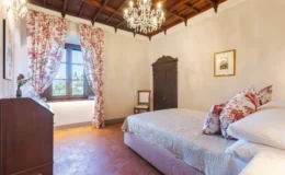 Cozy bedroom featuring a queen-sized bed with decorative pillows, elegant wooden furniture, and a vintage wardrobe. Natural light streams through large windows adorned with floral curtains, enhancing the inviting atmosphere. The room showcases a classic chandelier and terracotta tile flooring, perfect for a relaxing retreat.