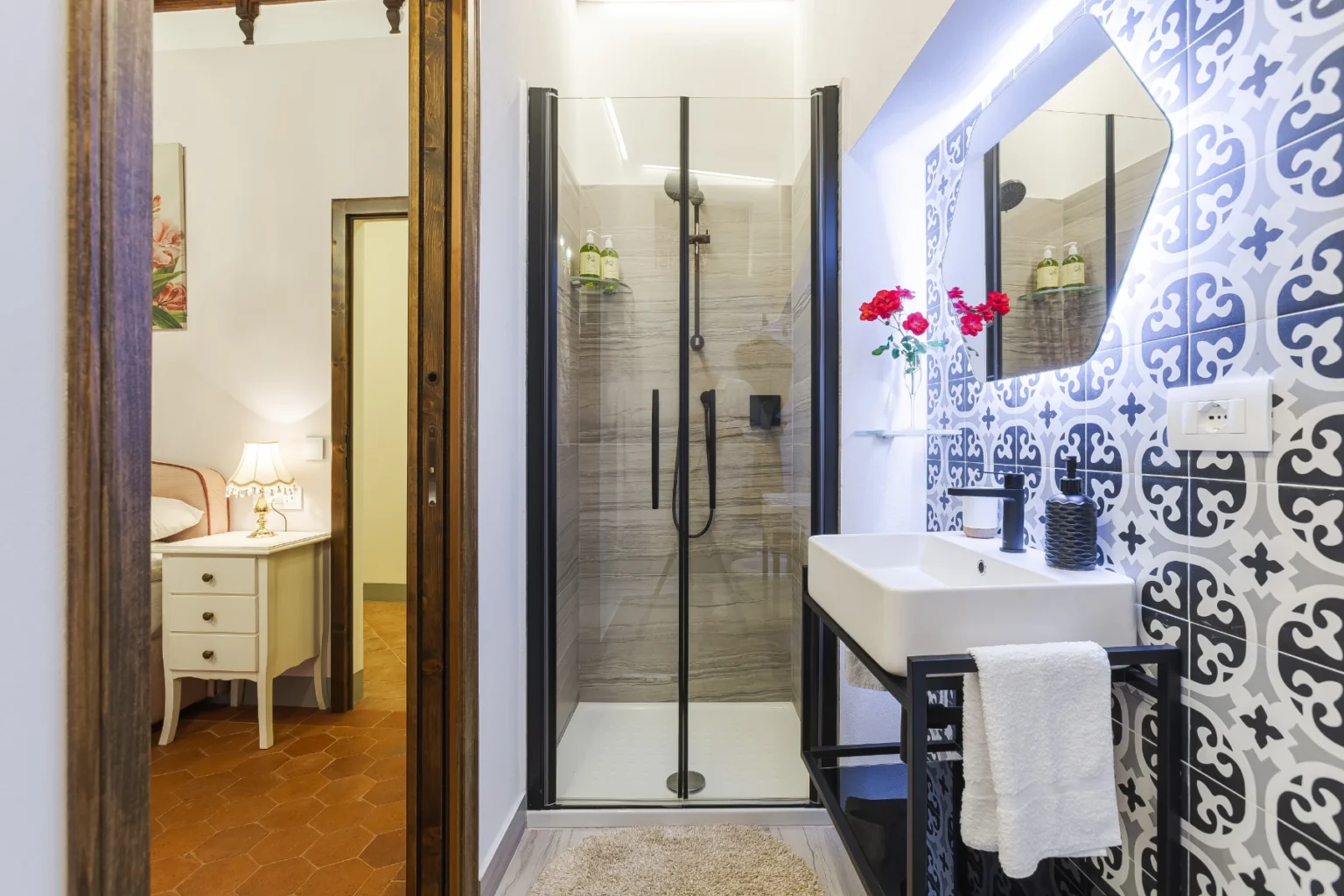 Modern bathroom featuring a glass shower enclosure, stylish sink with a black frame, decorative patterned tiles, and a mirror with integrated lighting. The space is accented with fresh flowers and has warm lighting from a bedside lamp in the adjacent room. | Only on SopranoVillas