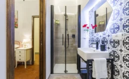 Modern bathroom featuring a glass shower enclosure, stylish sink with a black frame, decorative patterned tiles, and a mirror with integrated lighting. The space is accented with fresh flowers and has warm lighting from a bedside lamp in the adjacent room.