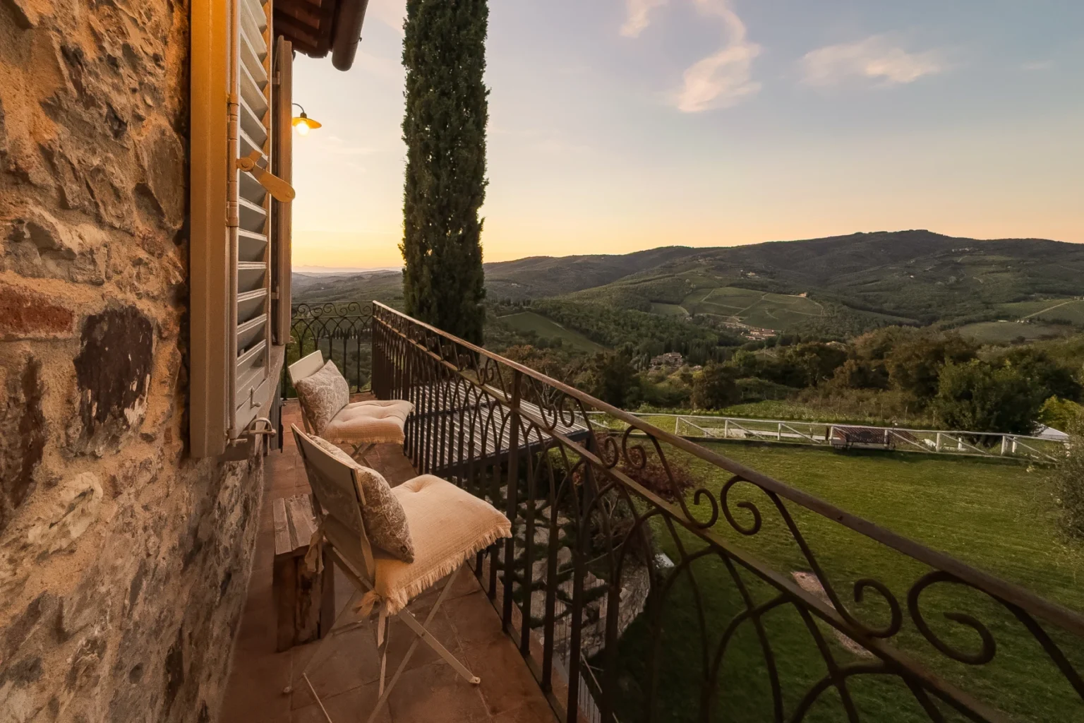 Scenic balcony view overlooking rolling hills and vineyards at sunset, featuring rustic stone walls, elegant seating, and lush greenery in a tranquil countryside setting. | Only on SopranoVillas