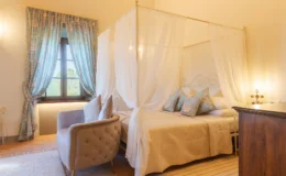 Cozy bedroom featuring a four-poster bed with sheer curtains, decorative pillows, and soft lighting. The room includes a plush armchair, patterned curtains, and warm-toned walls, creating an inviting and relaxing atmosphere.